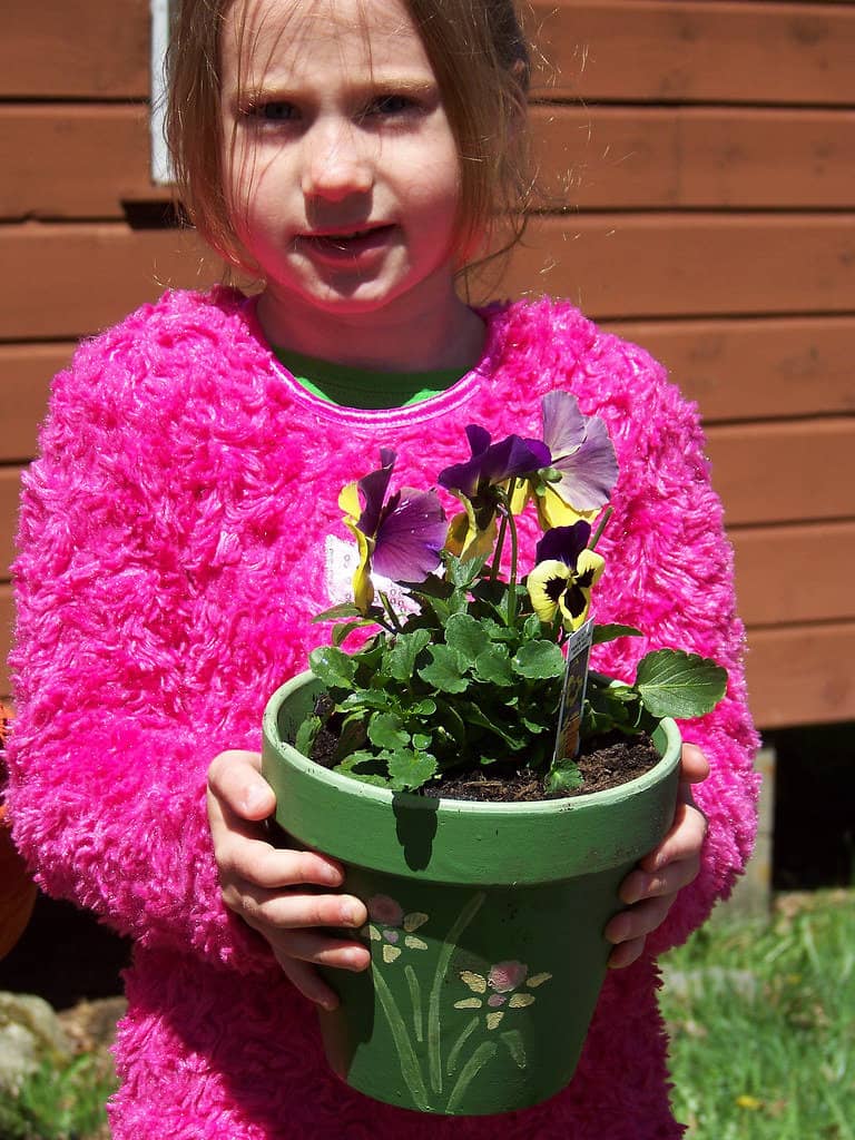 Adorable girl in a pink sweatshirt proudly holds a plant pot with a hand-painted floral design filled with flowers.
