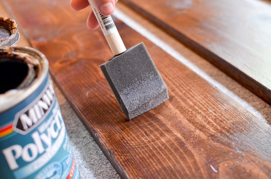 A hand holding a brush applies wood stain to a wooden surface.