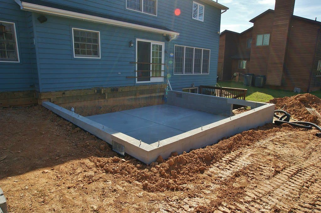 A foundation block for a shed has been laid down in the backyard of a blue house, surrounded by soil.