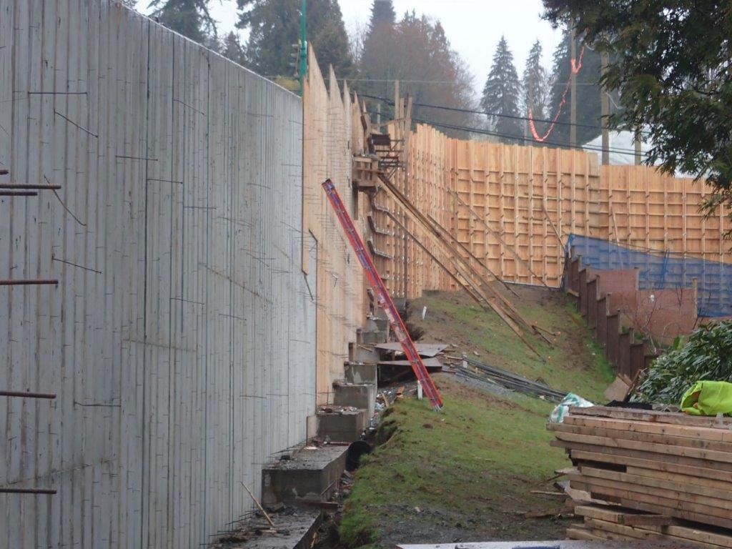 A concrete retaining wall is under construction on a sloped garden area, with wooden formwork supporting the structure.