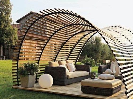 a half-cylinder on it's side with wooden plank cladding with gaps in between the planks to provide shade and sunlight at the same time, covering some garden furniture such as a sofa and a table in the middle of a garden 