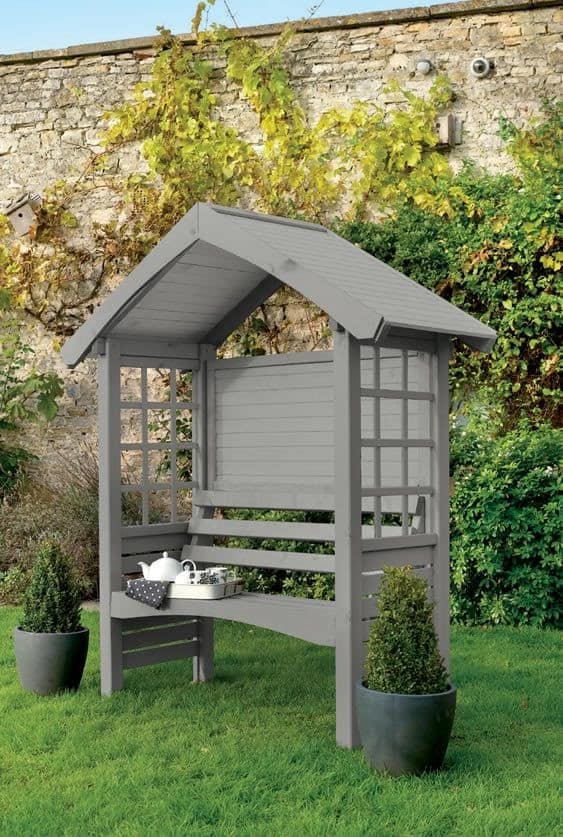 a grey bench in a little birds house type shelter with a roof and a wall at the back but no wall at the front