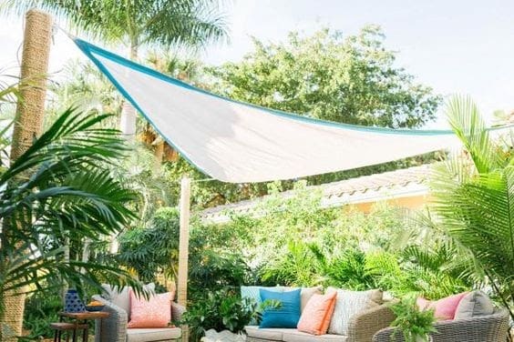 a white triangular sheet which is quite transparent allowing light in but reducing heat over some colourful cushions on an outdoor sofa in a tropical garden