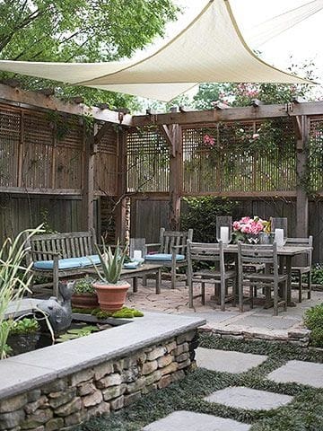 a dark wooden old looking trellis in a japanese-style garden with stepping stones and stone decking with some concrete groundwork and a table, chairs, plantpots and a beige cloth covering the area to create shade.