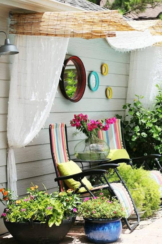 canopy covering two deck chairs in a garden with some colourful mirrors hanging from the wall above and covered by a pair of thin satin curtains held up by some wooden wood bamboo rolls.