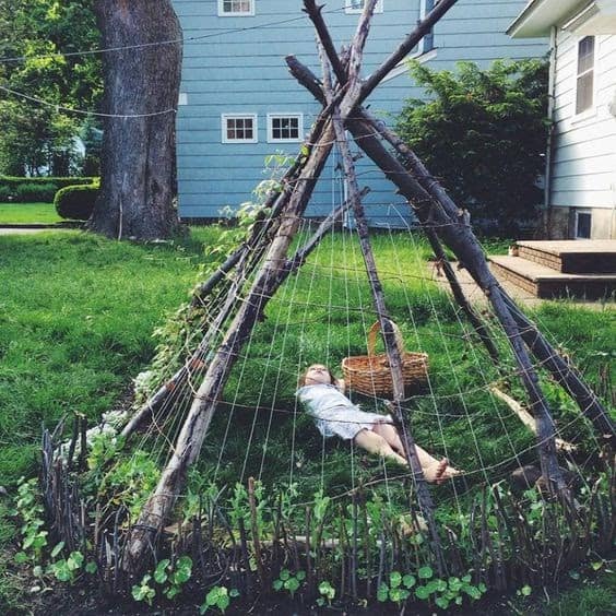 a teepee/wigwam made out of long wooden sticks from trees all slanted diagonally and meeting in the middle, with a child underneath with a woven basket. the teepee has spider web like strings woven around it to create a shelter and climbing plants climbing up it and a light blue house in the background and it is on some grass