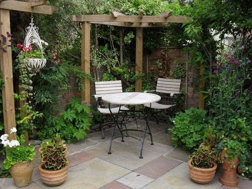 a corner shaped pergola made out of wooden beams set on some concrete patio with some garden table and chairs and lots of plants and greenery and a hanging basket.