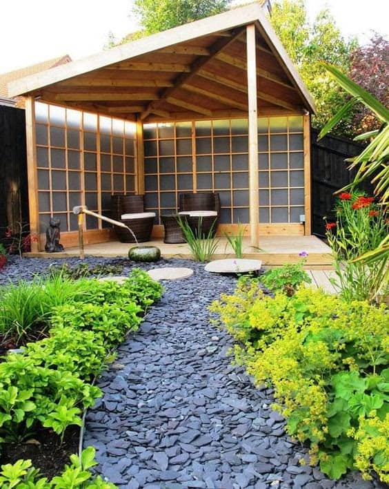 two comfy rattan chairs and a small table covered by an opaque triangular corner shaped wooden pergola held up by a single plank of wood at the tip of the triangle, with some translucent glass on the two walls and a shingle pathway leading up to it