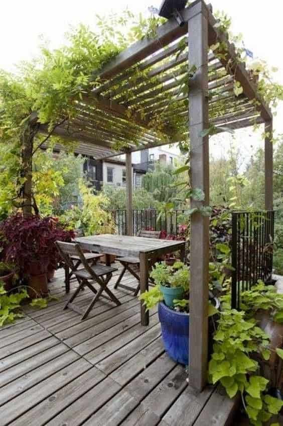 a wooden pergola/canopy with wooden beams spanning the roof, covered by climbing plants, moss and trees and on some decking with a table in the middle and some metal gating around the outside of the decking