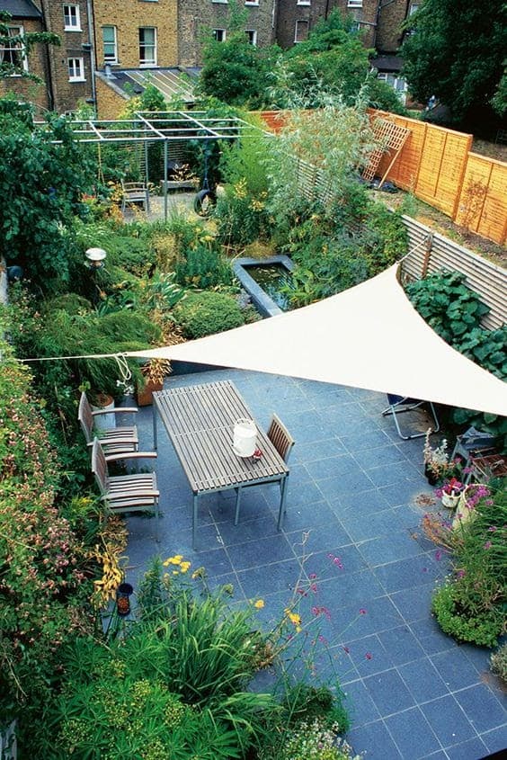 a triangular piece of cloth covering a blue patio with a table, chairs and furniture underneath in a floral garden with a pond and lots of leaves, plants and bushes 