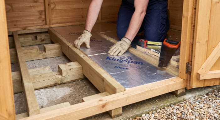 Close-up of a person installing foil-faced PIR boards within elevated wooden joists, demonstrating the necessary air gap and friction fit.