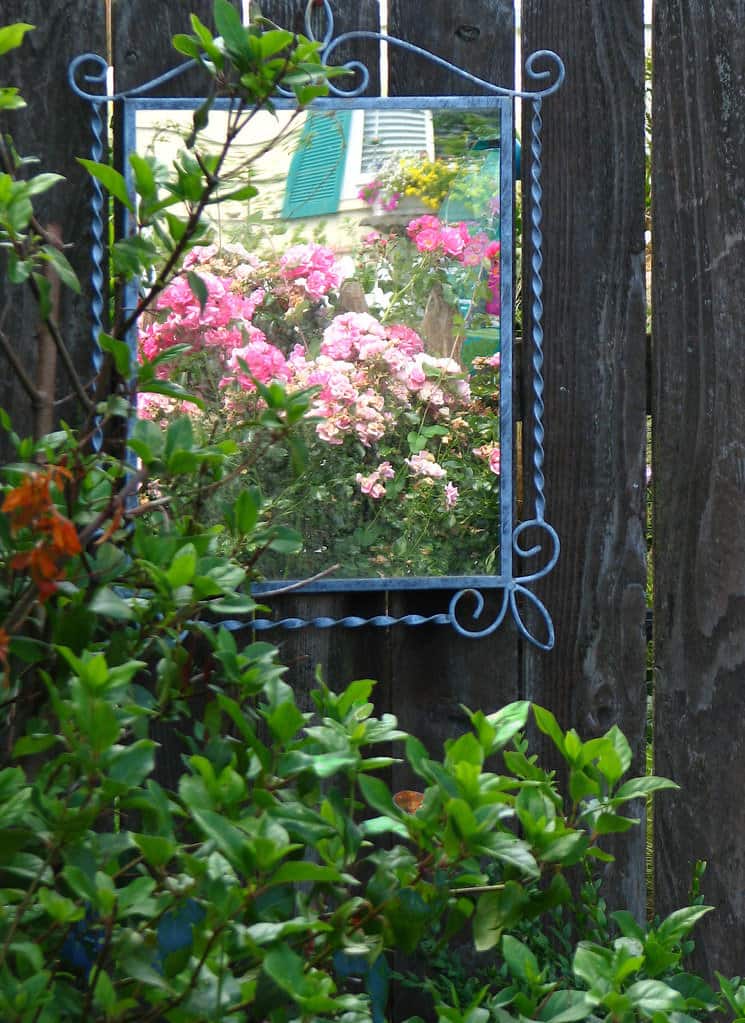 Blue-framed garden mirror on a wooden fence featuring artistic iron detailing.