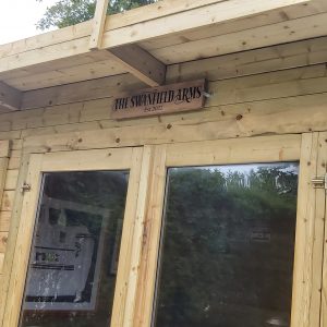 A sign reading "The Swanfield Arms" above the door of a log cabin