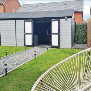 A large windowless shed painted grey with open double doors and a gravel path leading to it through a grass lawn.