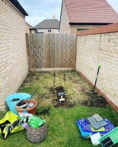 A section of grass garden, surrounded by fence and walls, that is being dug up with tools.