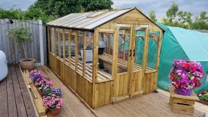 A wooden greenhouse surrounded by flower pots and beds in a wooden decked garden