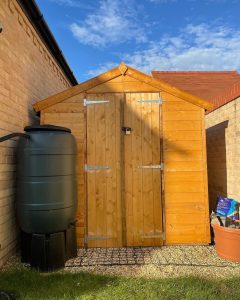 A shed on a gravel base with a water tank stood in front of it in the sunshine.