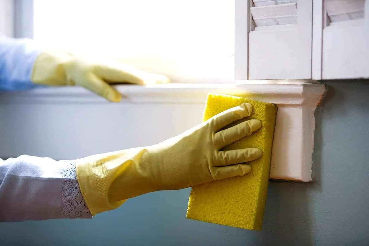 A person wearing yellow cleaning gloves is using a sponge to clean the window sills and frame