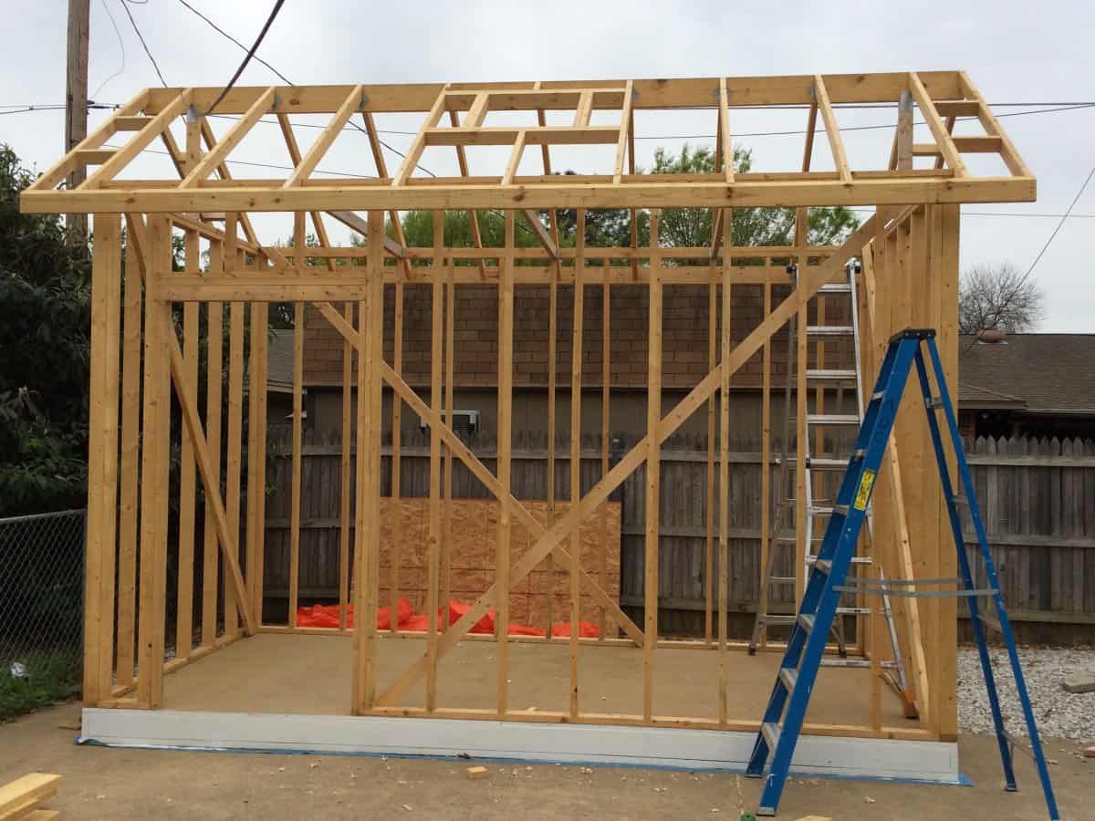 A wooden shed with beams and a ladder, set in an outdoor construction environment. The structure is partially completed.