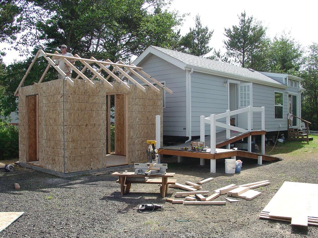 A nearly completed shed being built next to a house, with tools and equipment scattered on the ground.