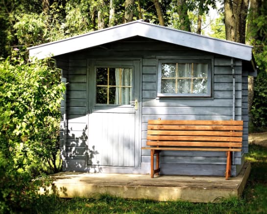 A mini garden shed painted in blue, with a wooden bench situated at front