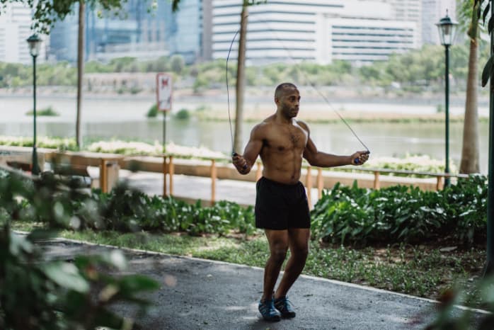 Topless man holding a jumping rope on the walkway of a park beside the lake.