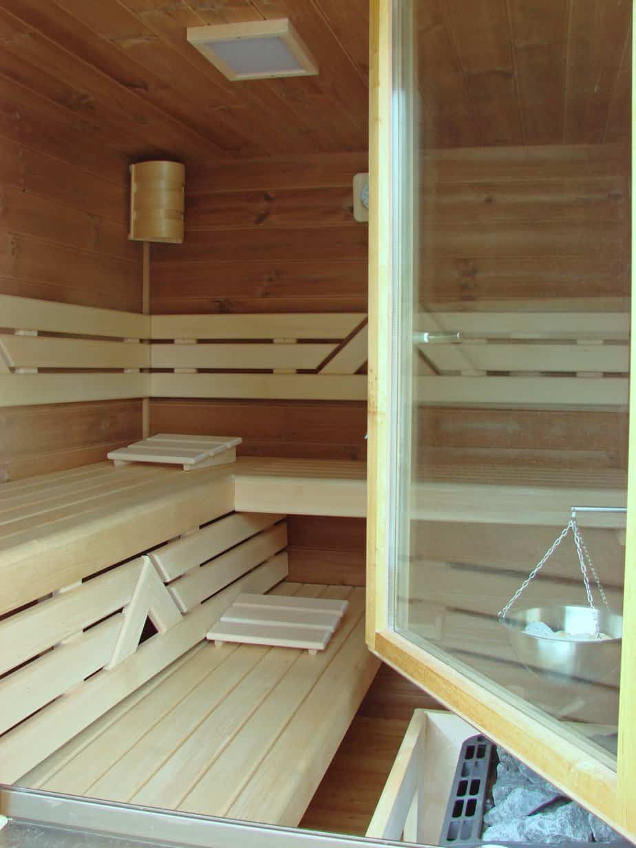 An interior of a wooden sauna, featuring light wooden benches and walls. A glass door is partially open, and a metal bowl on a chain hangs above the sauna heater filled with charcoals.