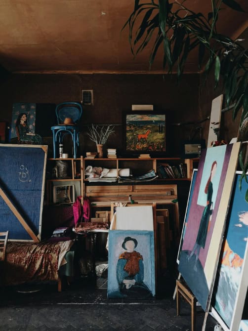 An artist’s studio filled with various paintings and art supplies - some are displayed on easels, and others are leaning against the wall.