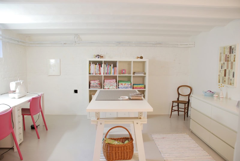 A bright sewing room with a white table, pink chairs, and a sewing machine. Shelves hold fabric and books, and a wicker basket sits on another table.