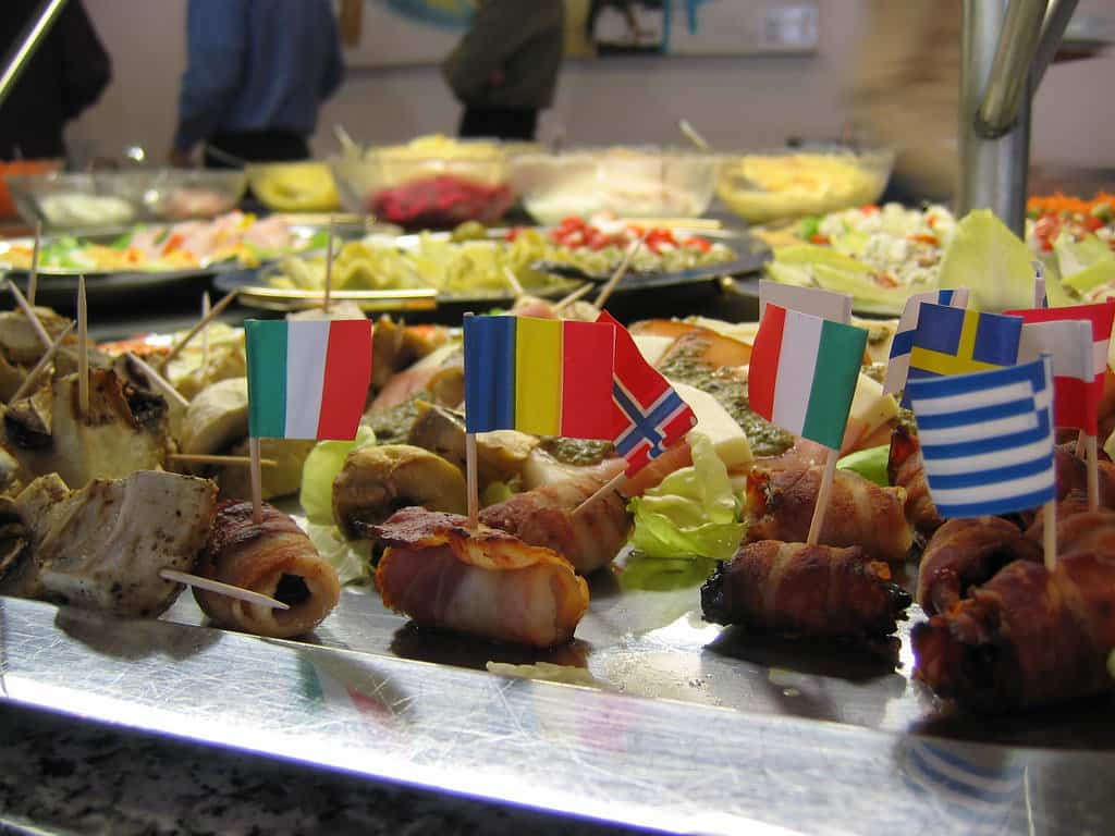 A buffet table with various appetizers, each adorned with small toothpick flags of different countries, including Italy, Romania, Norway, and Greece.
