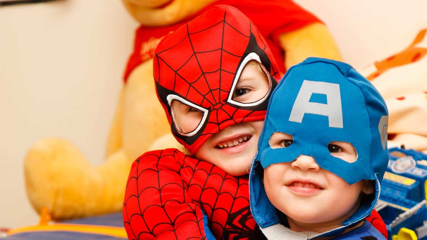 Two children dressed in superhero costumes, one as Spider-Man and the other as Captain America, smiling and hugging each other.