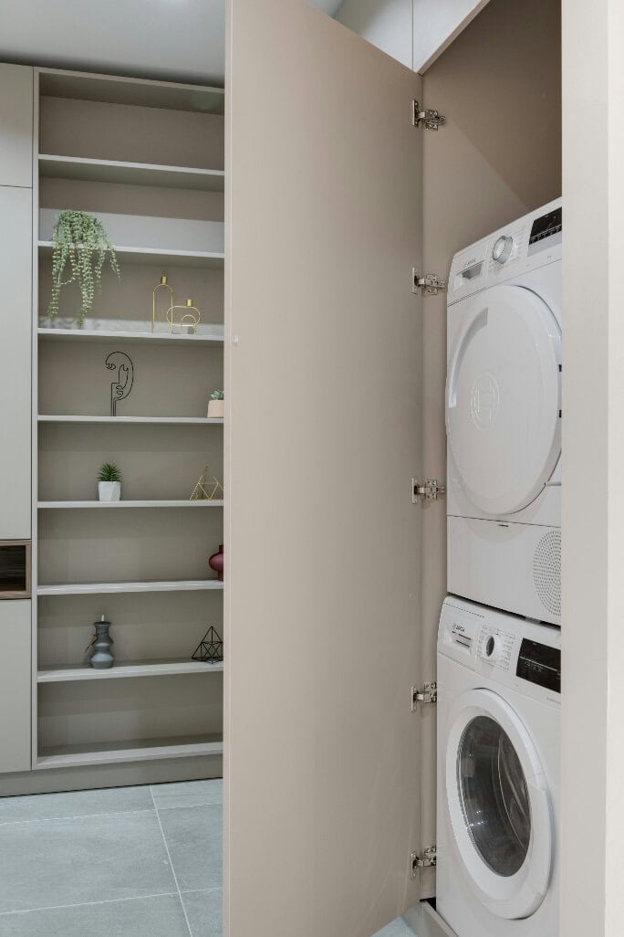 A laundry room with a stacked washer and dryer inside a cabinet, next to open shelves with decorative items.