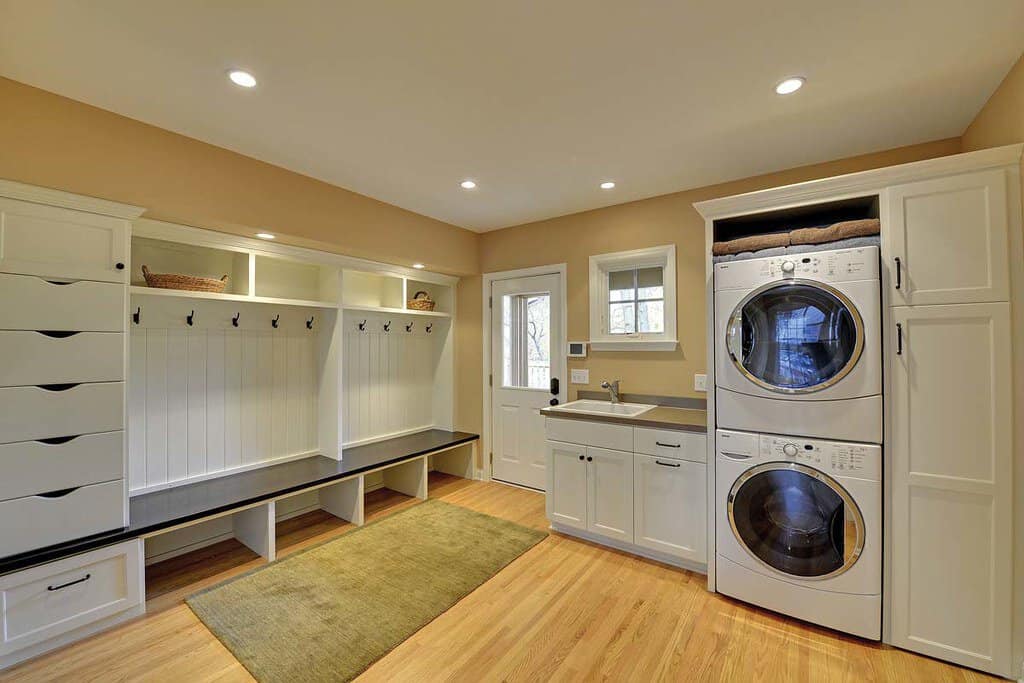 A spacious laundry room with stacked washer and dryer, a sink, built-in storage cabinets, and a bench with hooks.