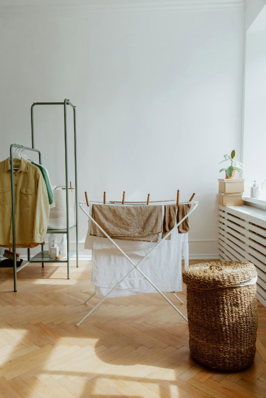 A laundry room with clothes drying on a rack, a woven basket, a plant on the windowsill, and a clothing rack.