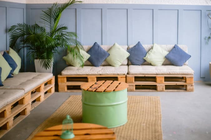 A patio corner with wooden pallet sofas adorned with green and blue cushions, a green drum table, and a large potted plant in between the L-shaped seating.