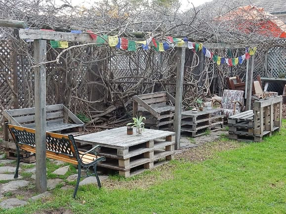  A garden area with a table made from stacked wooden pallets decorated with small potted plants. The table is part of a seating arrangement under a pergola with colourful flags.