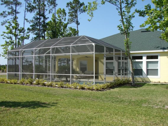 A sunroom with a metal frame and glass panels features floor-to-ceiling windows and a transparent roof. It’s attached to a yellow house and has an open view of the backyard.