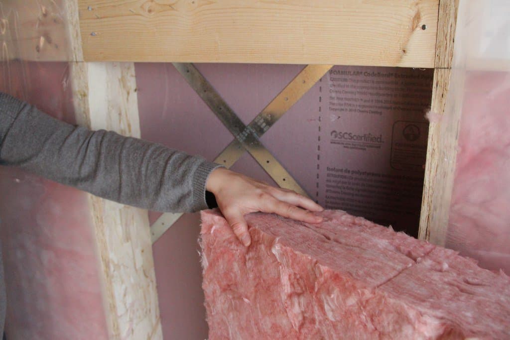 A person installing pink fibreglass insulation between wooden studs in a wall.