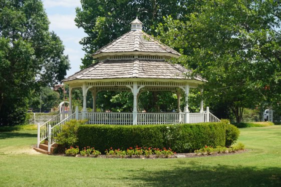 A gazebo situated on a solid foundation, surrounded by green grass and trees.