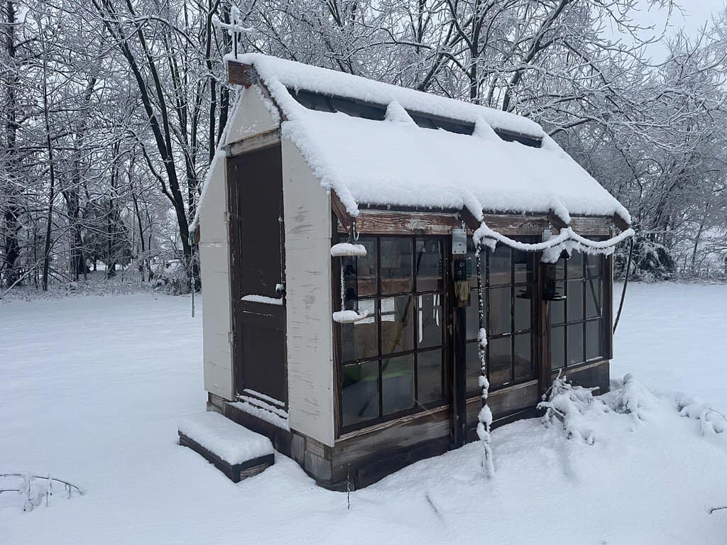 A greenhouse blanketed in snow, with snow accumulating on the roof.