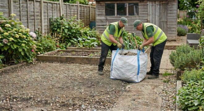 Two skip hire personnel in reflective vests and caps prepare a large woven bulk bag for filling.