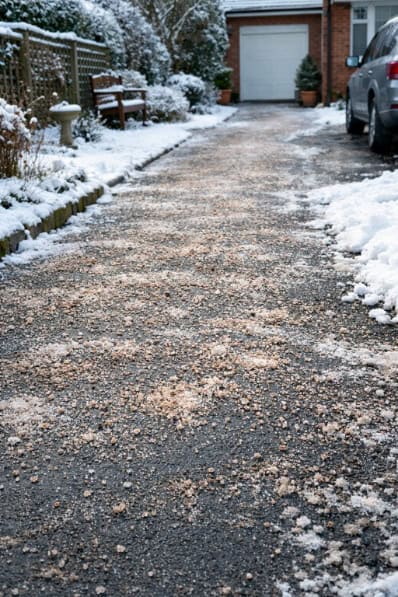 A home driveway and garden path in winter, lightly covered with salt grit and surrounded by snow.