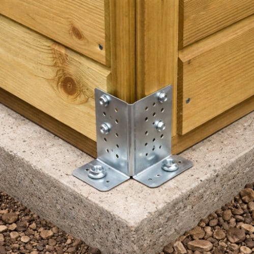 Close-up of a wooden garden shed corner secured to a concrete base using a metal corner bracket and bolts.