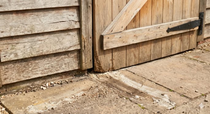A wooden shed door showing damage and wear at the bottom edge where it rubs against the paved ground.