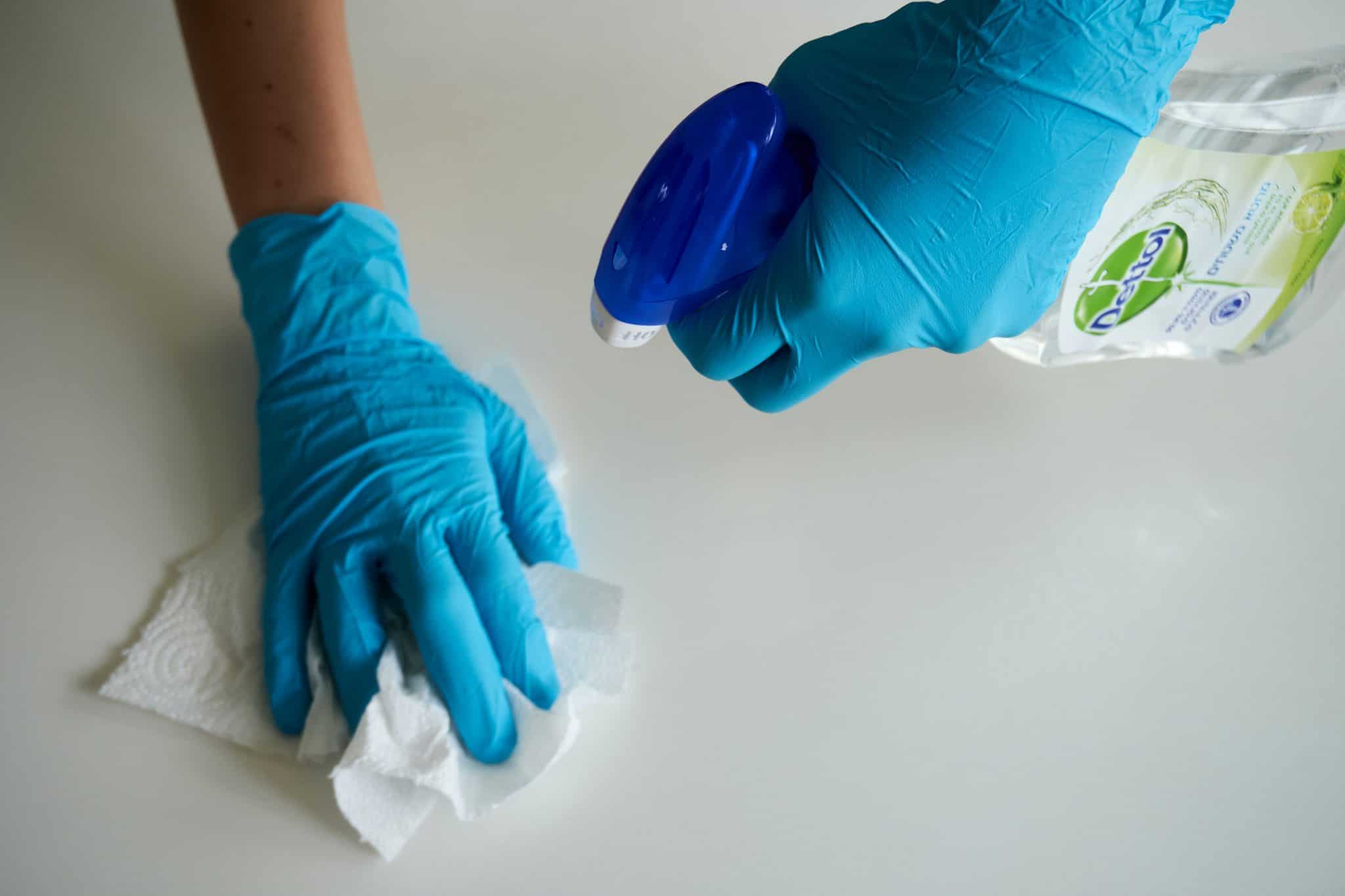 White surface with hands wearing blue plastic rubber gloves cleaning with a Dettol spray bottle and wipe