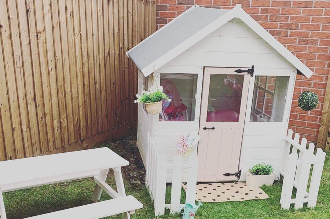 Single storey pent roof playhouse painted white with small bench and picket fence on turf