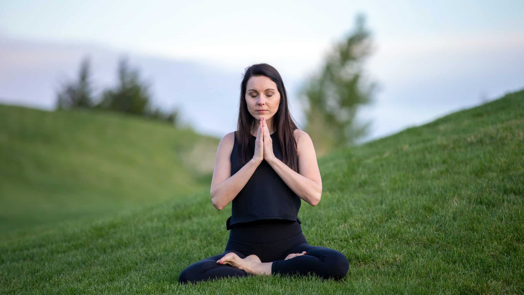 Woman sat cross legged with hands together in prayer on a green grass slope meditating