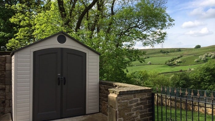 plastic black and white shed on a patio with a low brick wall backed by trees and fields