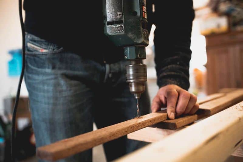 man wearing jeans drilling a hole in thin length of timber